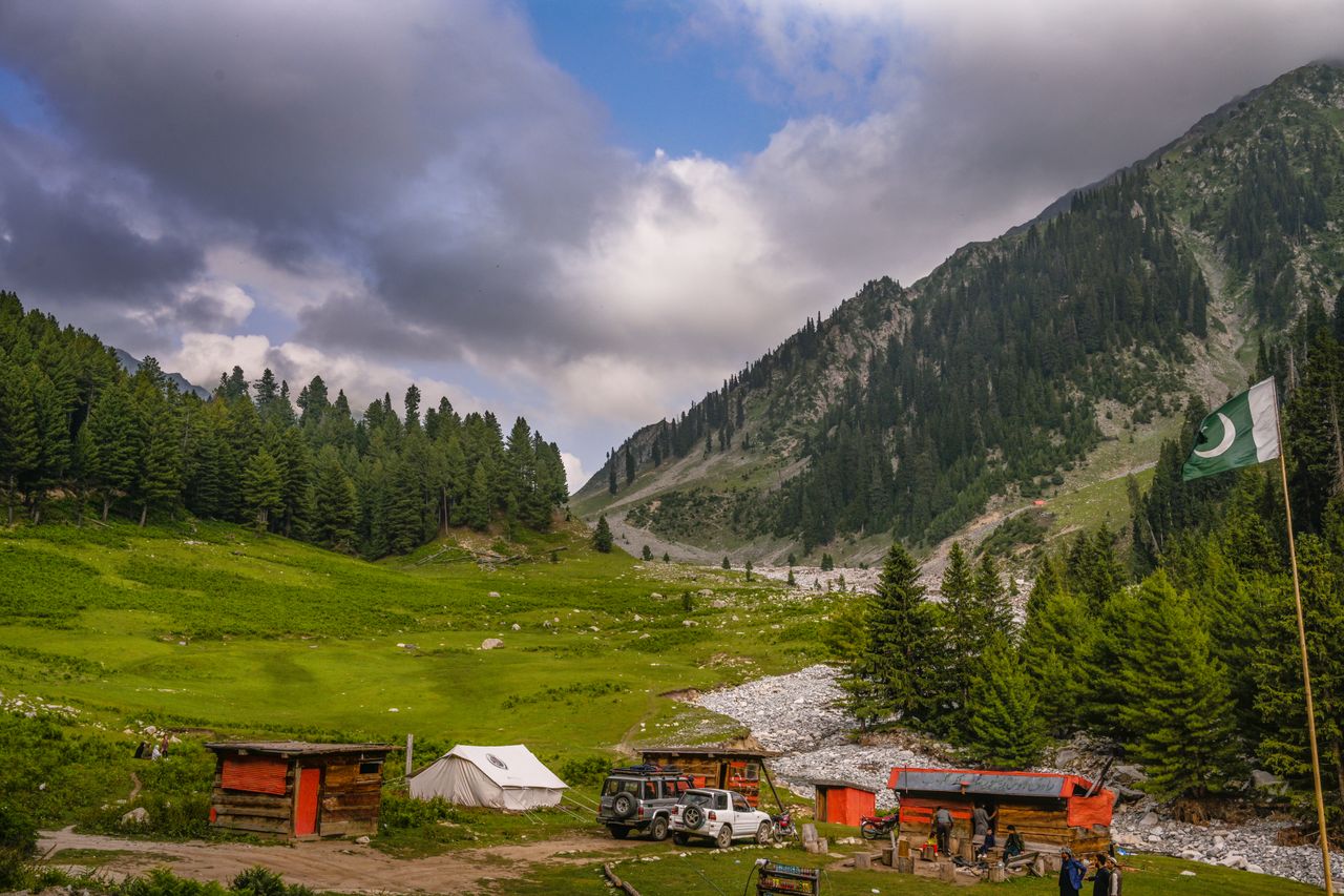 our-story Lush green valley with mountains, a few huts, and a national flag under a cloudy sky.